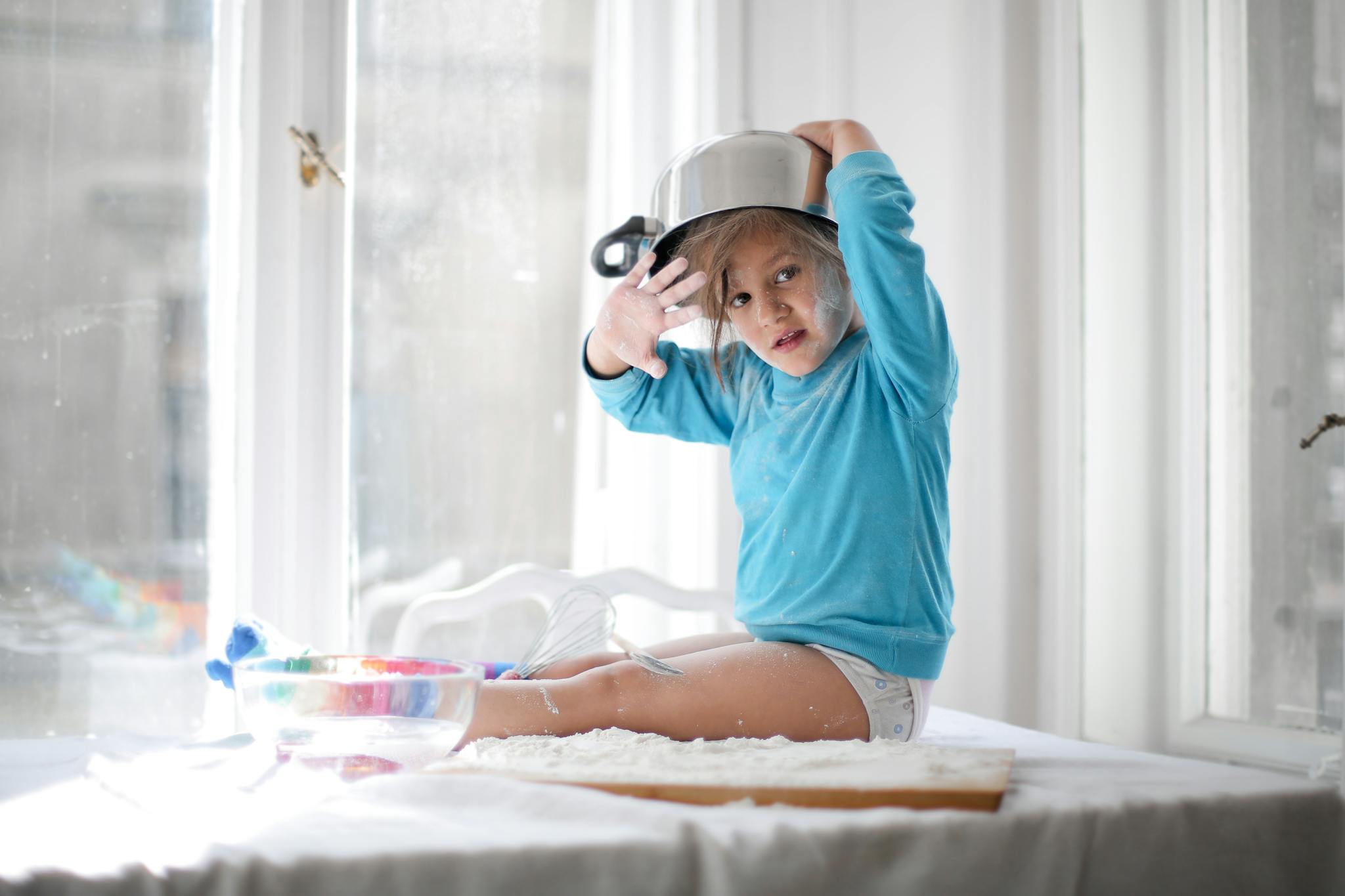 A child in a blue sweater playfully wears a pot on their head while sitting on a kitchen table.