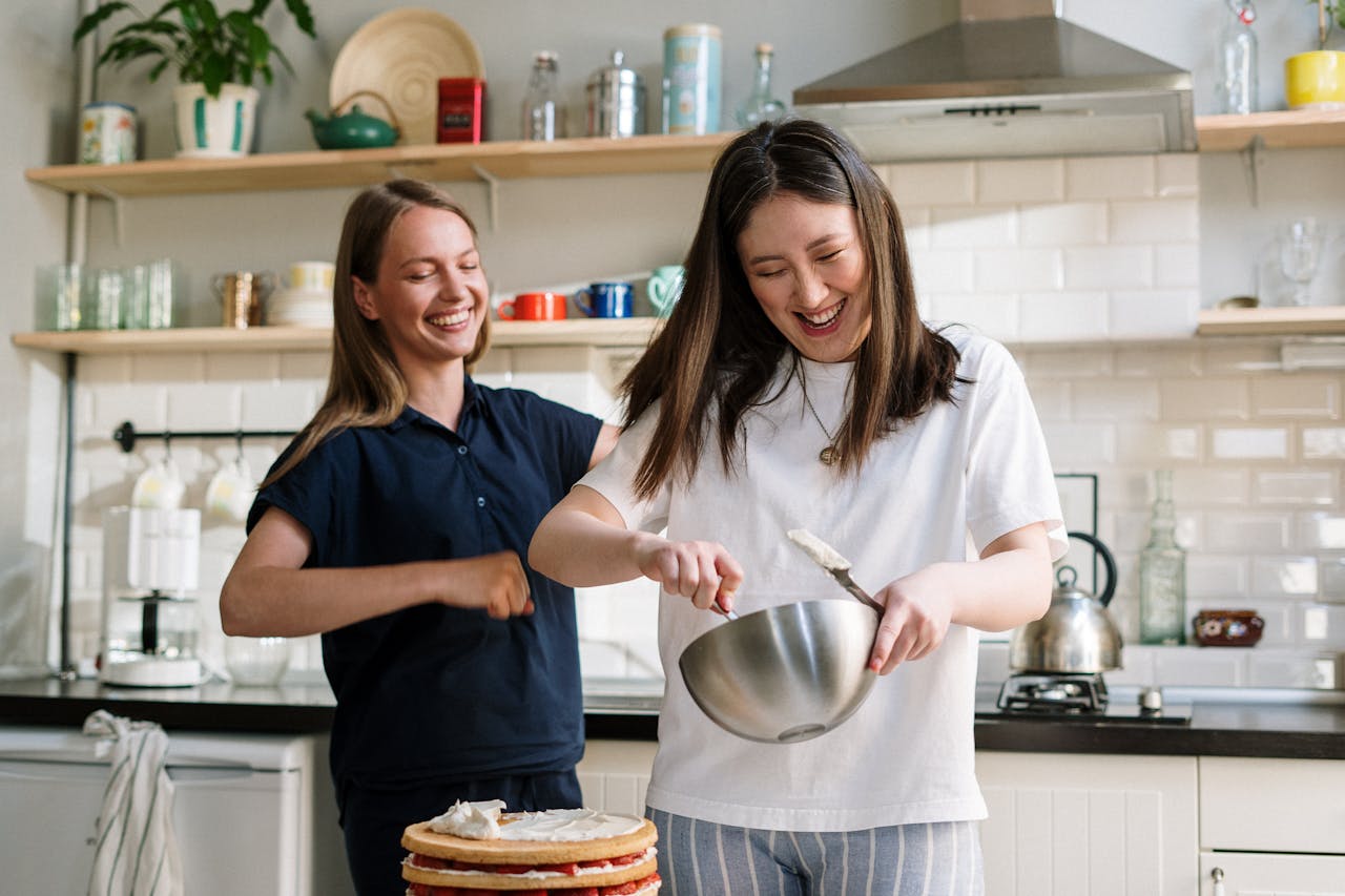 Two women cooking and laughing while preparing a cake in a cozy kitchen.